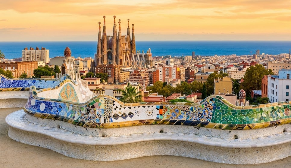 A panoramic sunset view of the Barcelona skyline from Park Guell, featuring the colorful mosaic serpentine bench in the foreground and the spires of the Sagrada Familia in the distance.